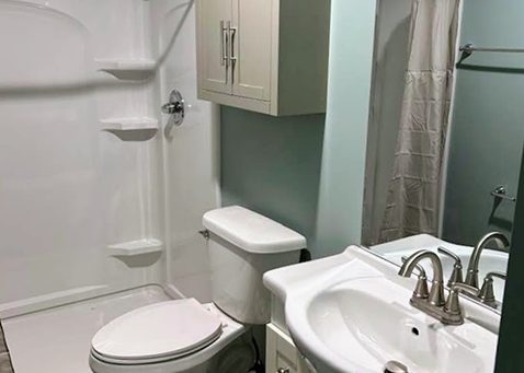 A small, bright bathroom with a white shower, toilet, and vanity sink, featuring light blue walls and a wall-mounted cabinet.