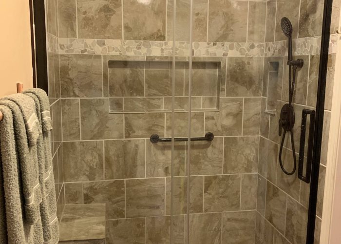 Walk-in shower with earth-toned tile walls, pebble stone floor, glass sliding doors, built-in bench, and rainfall plus handheld showerheads.