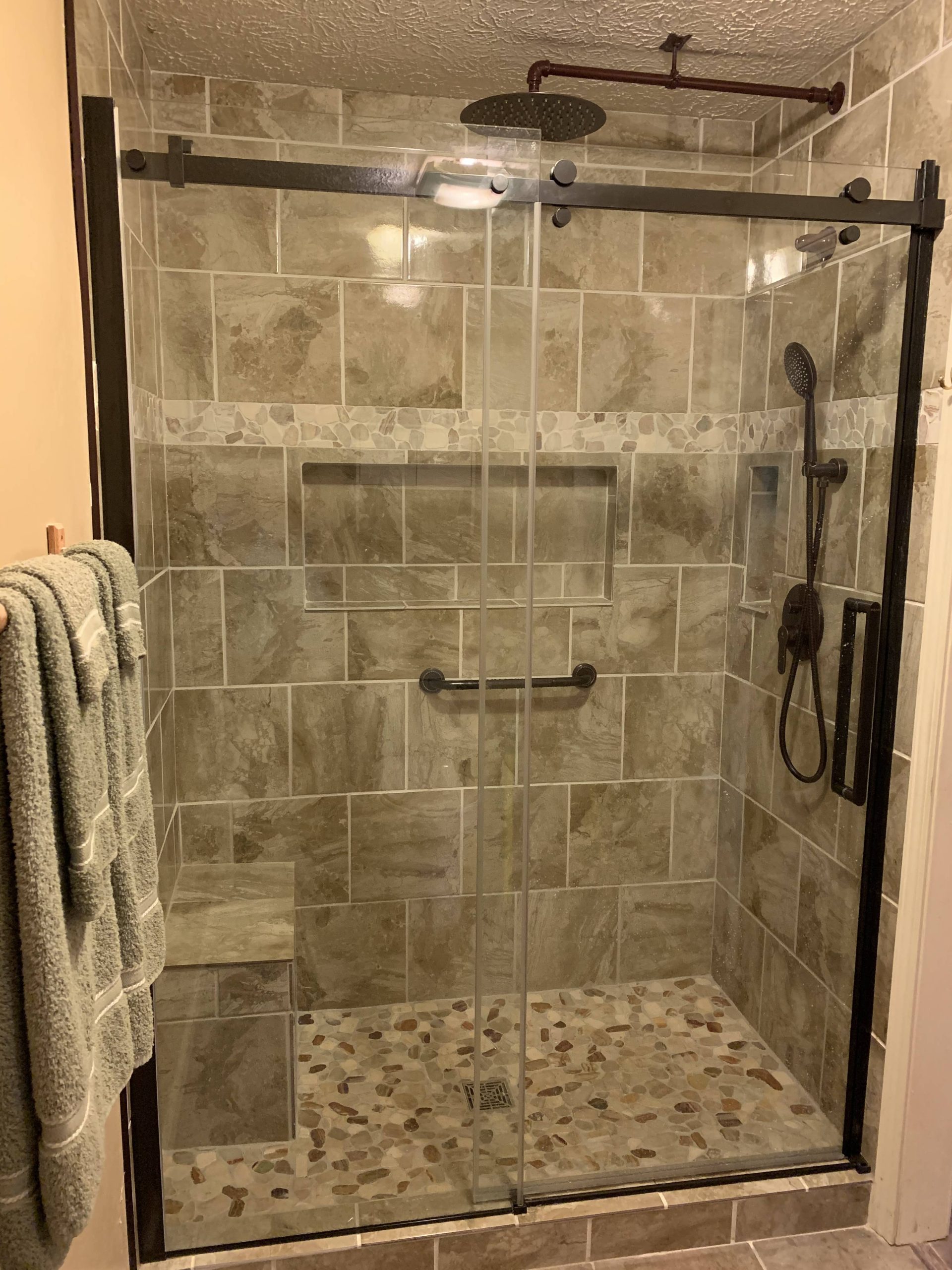 Walk-in shower with earth-toned tile walls, pebble stone floor, glass sliding doors, built-in bench, and rainfall plus handheld showerheads.