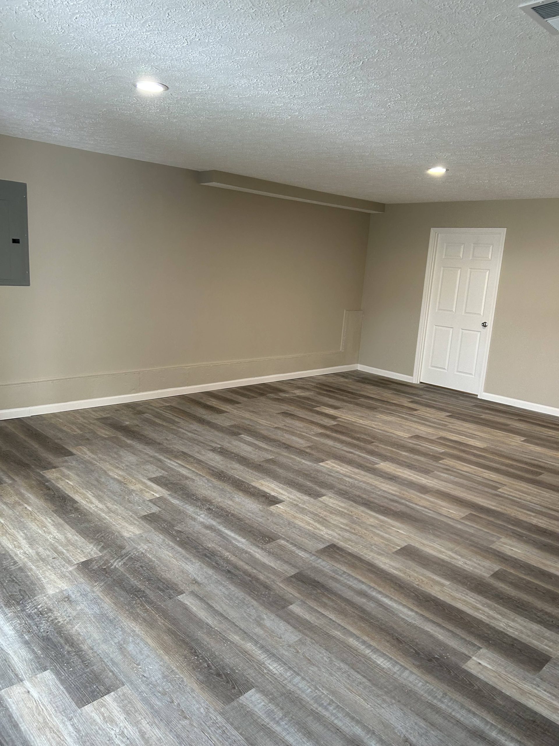 Finished basement room with gray-brown vinyl plank flooring, beige walls, white trim, and recessed ceiling lights.