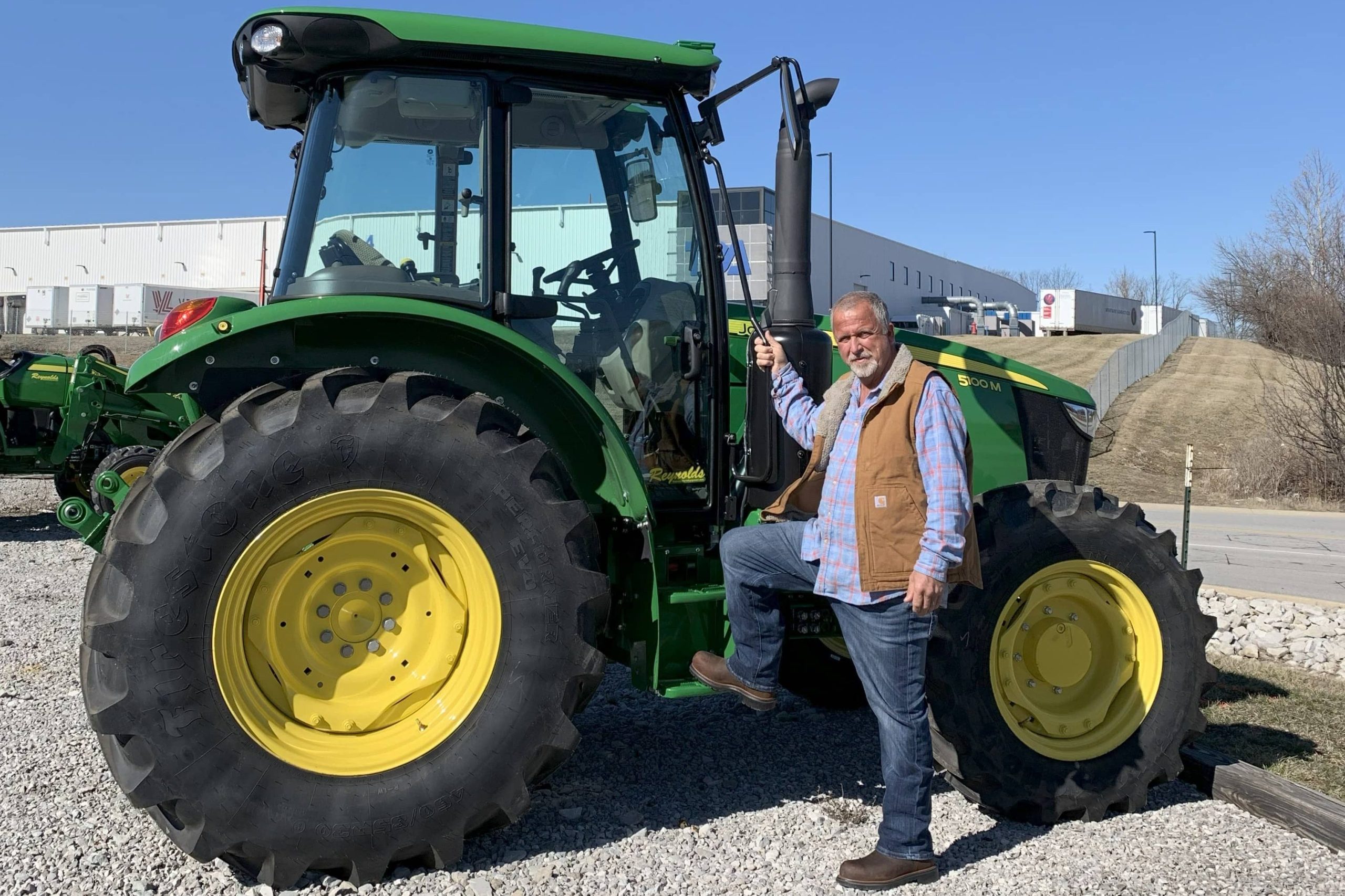 Joe Workman standing next to a green John Deere tractor with yellow wheels on a gravel lot under a clear blue sky.