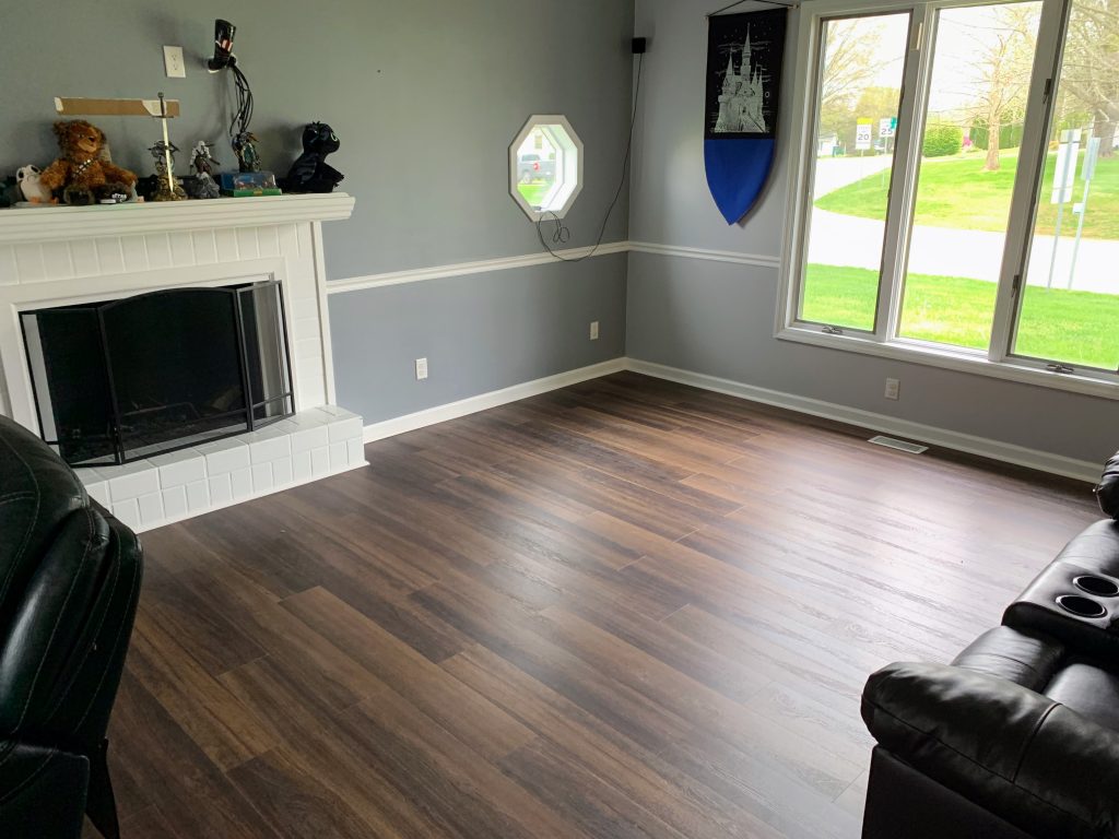 Living room with dark wood flooring, white brick fireplace, gray walls with chair rail trim, and large windows overlooking a green lawn