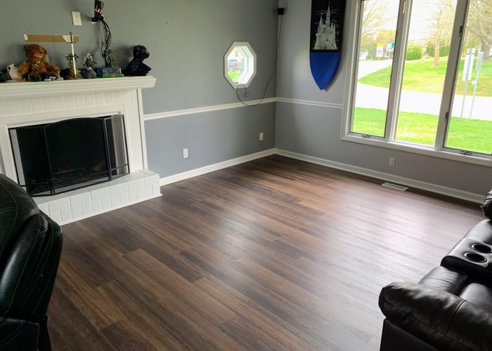 Living room with dark wood flooring, white brick fireplace, gray walls with chair rail trim, and large windows overlooking a green lawn