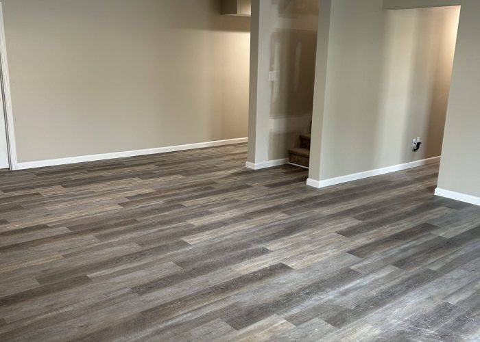Spacious finished basement with gray-brown vinyl plank flooring, beige walls, white trim, and a partially finished stairway area.
