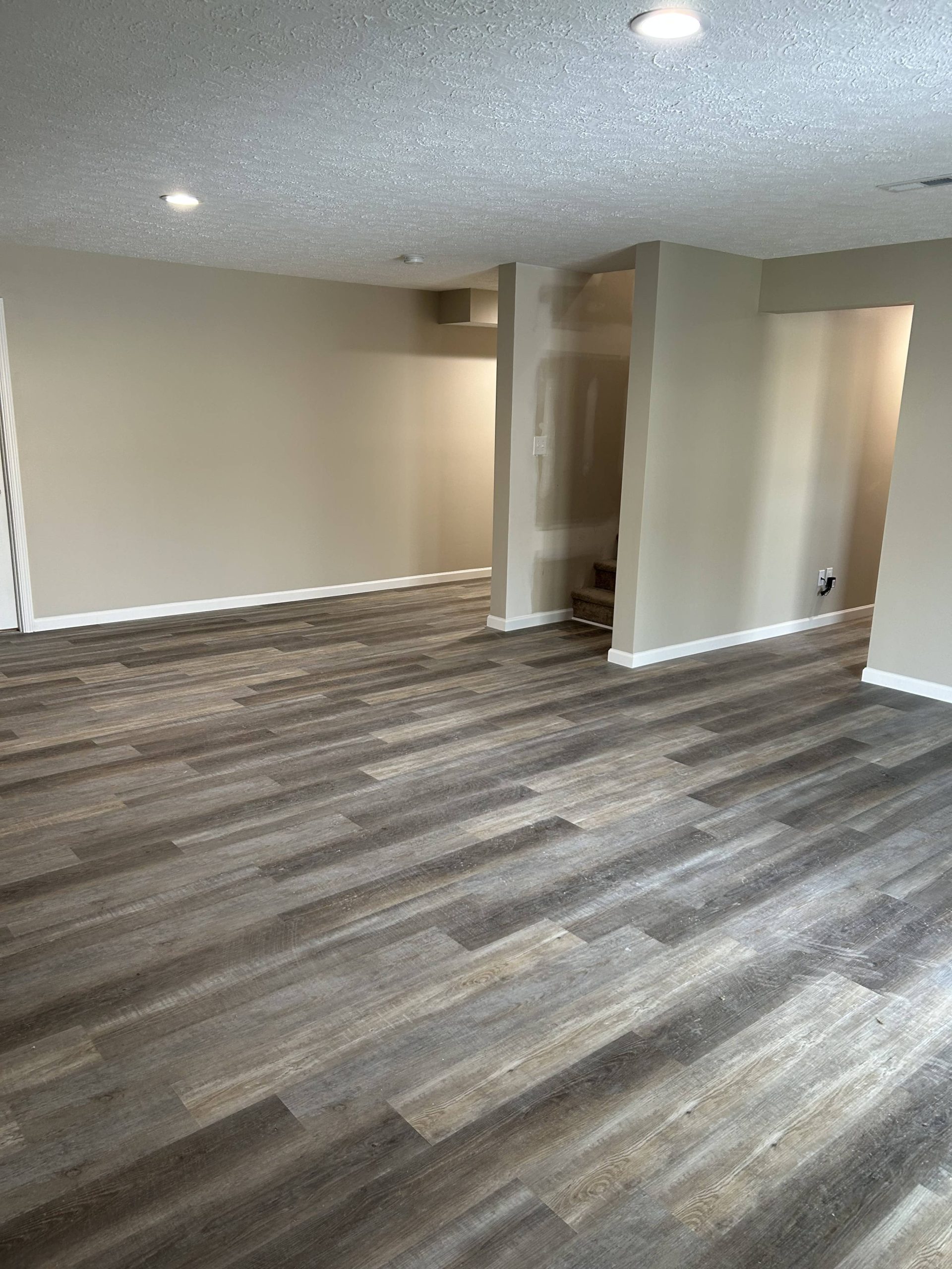 Spacious finished basement with gray-brown vinyl plank flooring, beige walls, white trim, and a partially finished stairway area.