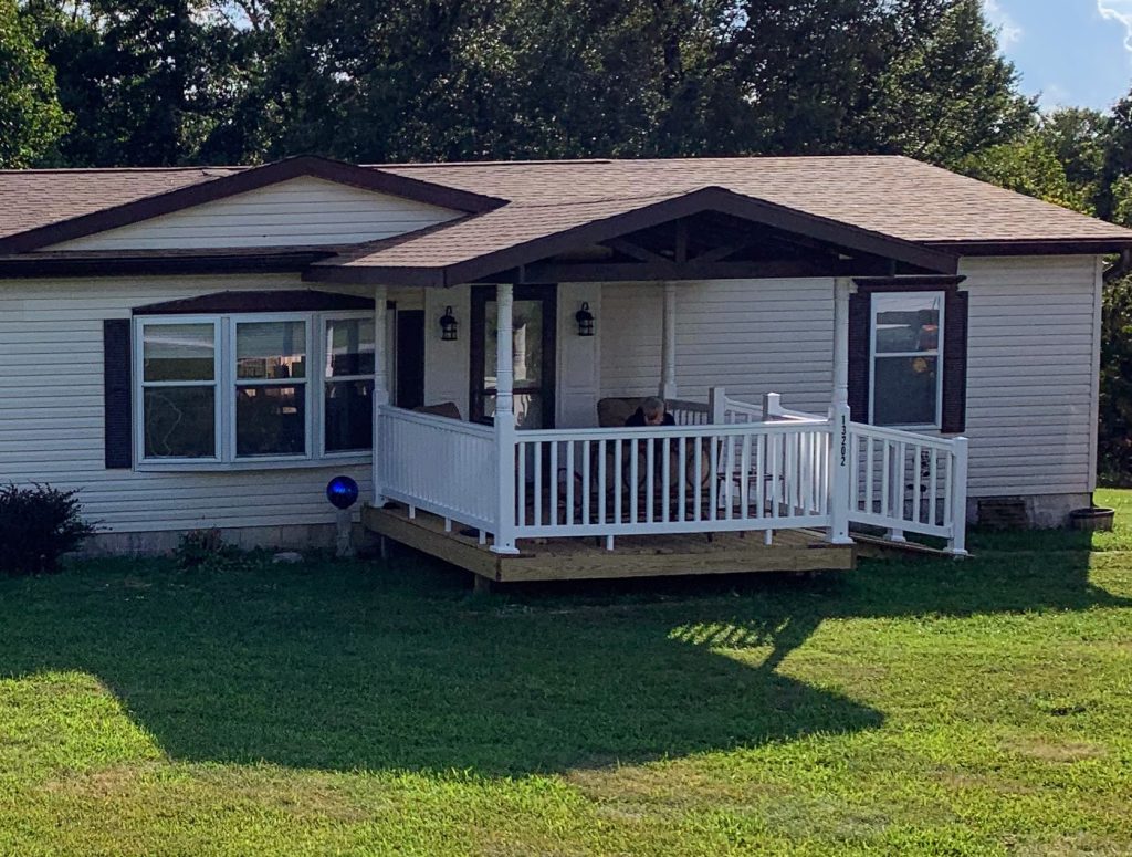 Single-story white manufactured home with a gable roof, covered front porch, and white railing, set on a grassy lawn with trees in the background