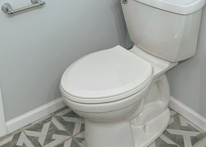 White two-piece toilet installed in a light gray bathroom with geometric patterned tile flooring and a chrome toilet paper holder.