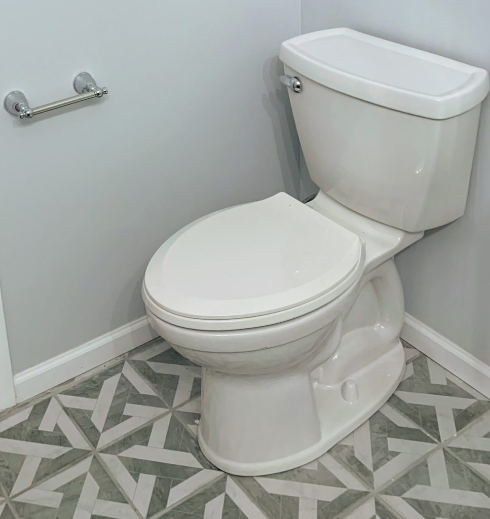 White two-piece toilet installed in a light gray bathroom with geometric patterned tile flooring and a chrome toilet paper holder.