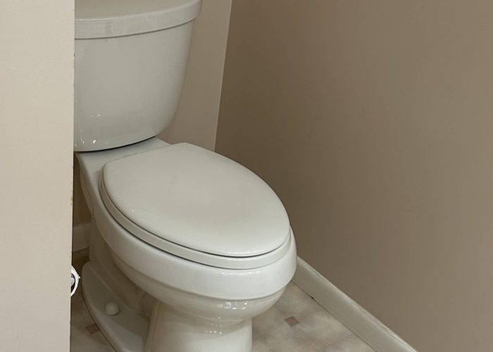 White two-piece toilet installed in a small bathroom corner with beige walls and patterned vinyl flooring.